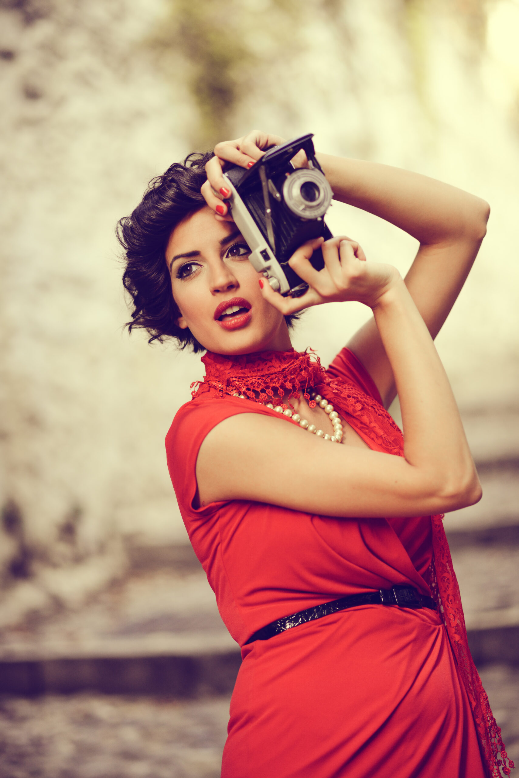 Portrait of a pretty woman, vintage style, in urban background, wearing a red dress taking photographs with a old camera